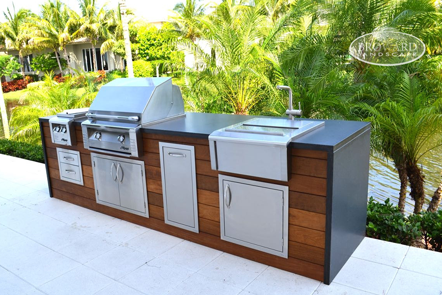Outdoor kitchen setup with grill, sink, and storage cabinets surrounded by greenery.